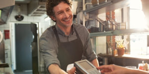 Waiter standing in a small restaurant smiling and handing a credit card payment scanner to a customer whose hands appear in the shot