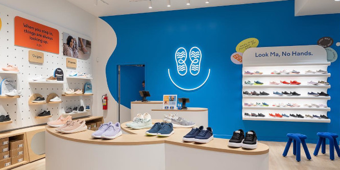 Image of the inside of Kizik's first physical retail location, shoes lined up on a table with a curving, modernist design in front of a blue wall with a while shelf built directly into it, above the shelf appear the words "look ma, no hands"