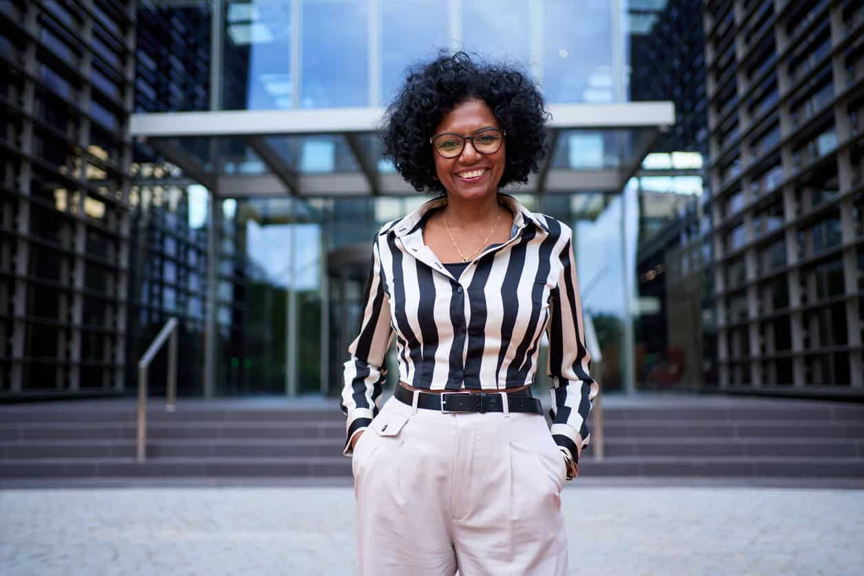 Modern African business adult woman standing hands in pockets looking joyful at camera outdoor.